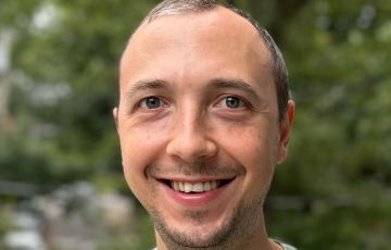 Robert, young white male with receeding hairline and a stubble mustache and beard wearing a green tshirt standing in front of blurred trees