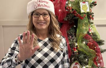 Lori middle aged white woman with long blond hair wearing a Merry Christmas santa hat and glasses and black and white geometric blouse standing in front of a Christmas tree