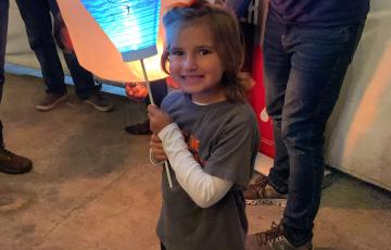 young smiling white girl in a gray shirt over a white long sleeved shirt with and orange bow in her hair and holding a LTN balloon