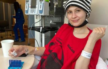young white woman wearing a black and white knit cap and a red t-shirt and black short with painted white fingernails laying in an infusion chair