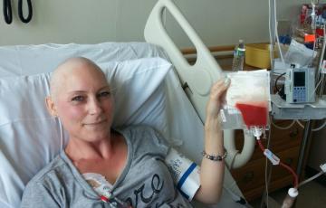 Babette young white woman who is bald waring a grey t-shirt and lying in a hospital bed holding an infusion bag