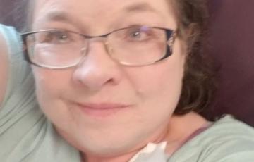 Dixie older white woman with brown hair and glasses wearing a green shirt laying against a brown pillow with an earbud laying on her chest