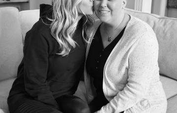 Janie young white woman who is bald wearing a black shirt and a white speckled cardigan being kissed by a young white woman with long blond hair wearing a black hoodie both sitting on a couch