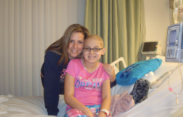 Kendra woman and young hispanic girl on a hospital bed girl has glasses and wearing a pink shirt and plaid pajama bottoms with turqouise pillow on bed