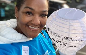 Kenya middle aged black woman with short black hair wearing big hoop earrings and a blue survivor tshirt and white ribbon hold a LTN light