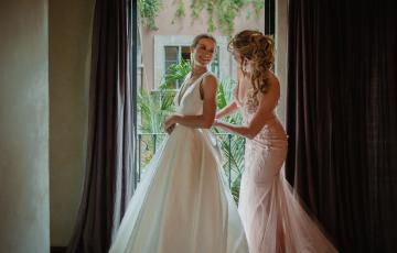 Laura young white woman in a white wedding dressing looking at her mother who has a curly hairdo and wearing a pink formal dress both standing in front of a balcony