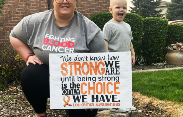 Maverick young white mother with a bun and glasses wearing gray blood cancer shirt holding a leukemia sign with her young son next to her in a gray shirt and smiling
