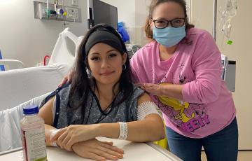Mayra young hispanci woman sitting in a hospital bed wearing a black headbank and blue hospital gown and has three face piercings with another woman with face mask and pink shirt next to her