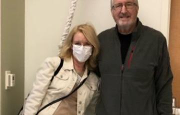 Rex older white couple standing in a hospital room man has white hair and glasses wearing a gray jacket and pajama pants woman has face mask