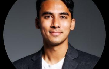 Rohan young man of Indian heritage with dark hair and slight chin stubble wearing a dark gray jacket over a white tshirt.